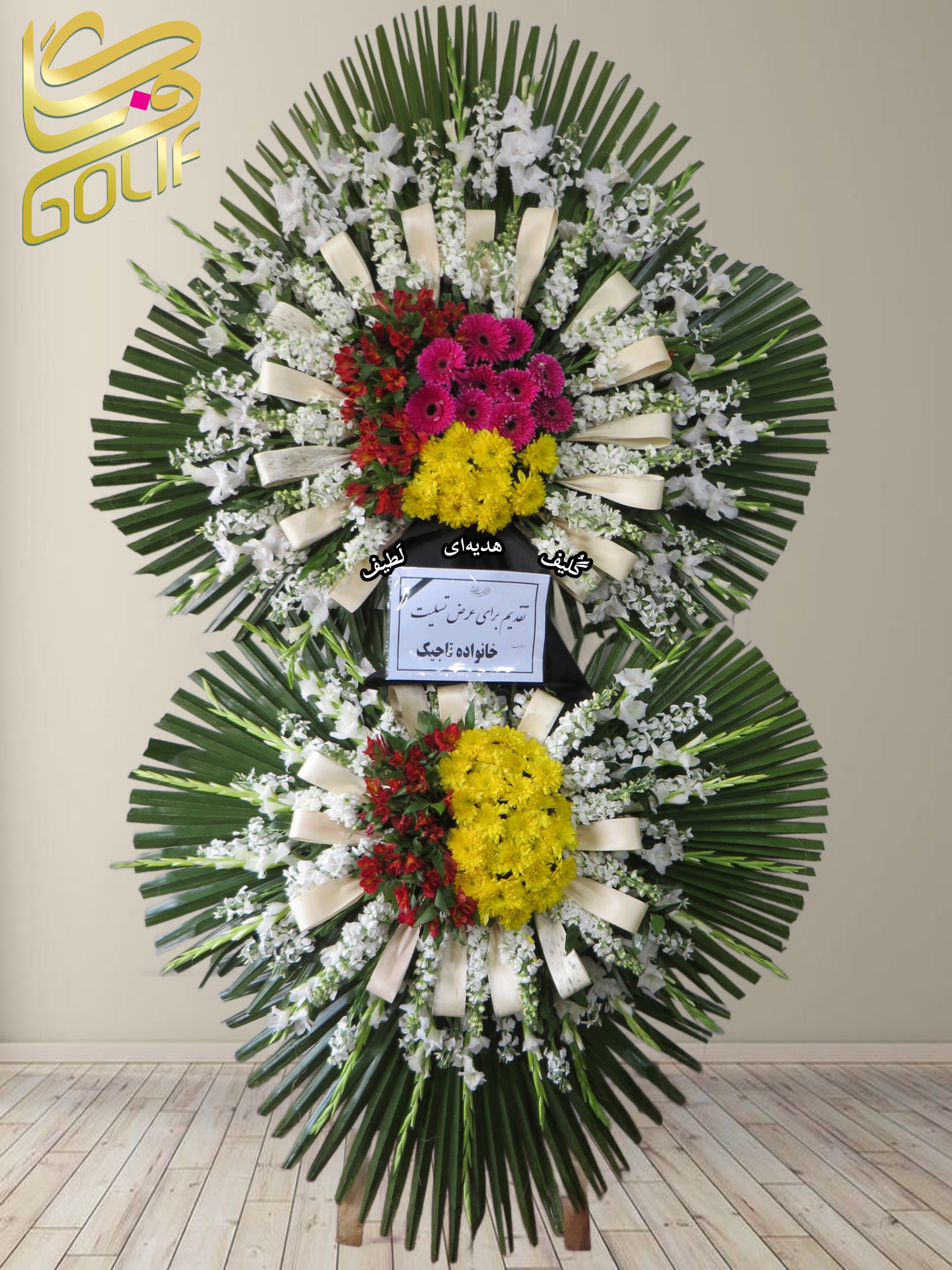 تاج گل تسلیت دو طبقه (لیلیوم، رز، آنتوریوم، داوودی) تاج گل ختم، تاج گل ترحیم، تاج گل مراسم ترحیم گل‌هایی که در این تاج گل دیده می‌شوند شامل موارد زیر هستند: لیلیوم: گل‌های زرد رنگ بزرگ و خوشبو که در قسمت بالای دسته گل حضور دارند. رز صورتی و قرمز: این گل‌ها با رنگ‌های جذاب در طراحی زیبای دسته گل استفاده شده‌اند. آنتوریوم: گل‌های قرمز و سفید که به طراحی دسته گل جلوه ویژه‌ای داده‌اند. داوودی: گل‌های زرد و سفید که معمولاً برای مراسم‌های مختلف از جمله تسلیت به کار می‌روند. علاوه بر این گل‌ها، برگ‌های سبز و روبان‌های کرم رنگ نیز استفاده شده‌اند تا دسته گل زیباتر و دلپذیرتر شود. سفارش آنلاین تاج گل سفارش آنلاین گل عکاسی شده توسط گل‌فروشی آنلاین گلیف