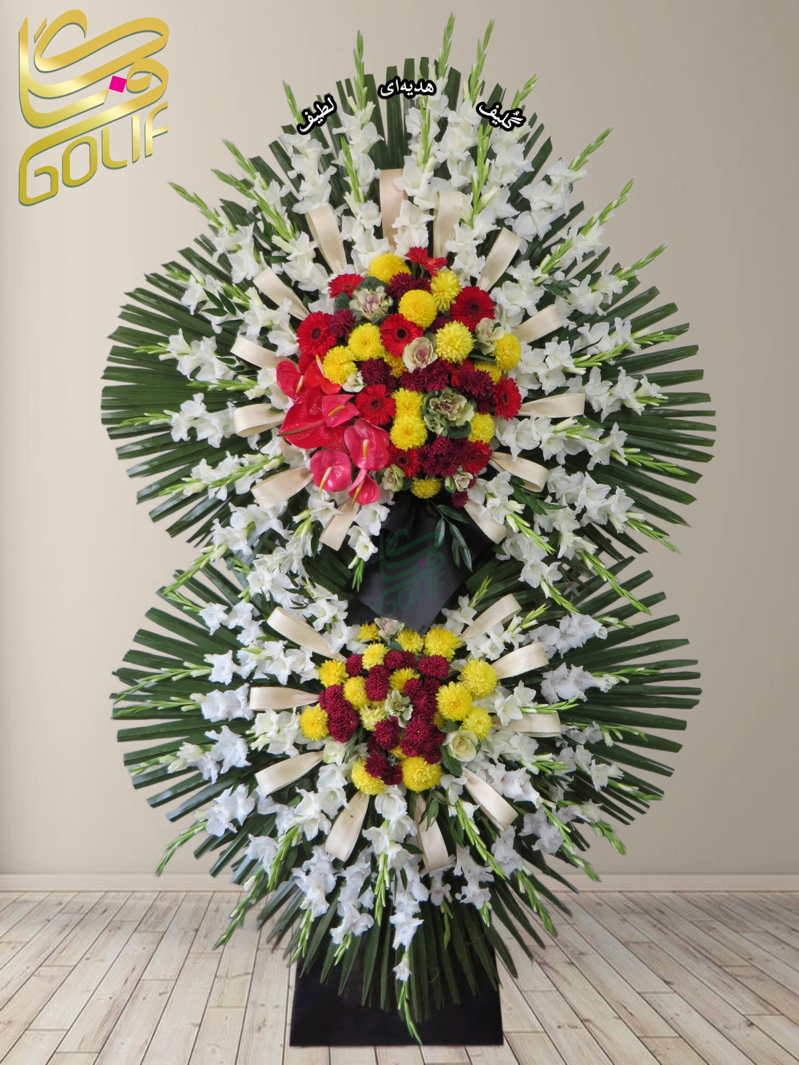 تاج گل تسلیت دو طبقه (گلایل، داوودی، ژربرا، آنتوریوم) تاج گل ختم، تاج گل ترحیم، تاج گل مراسم ترحیم این تاج گل تسلیت از ترکیبی چشم‌نواز از گل‌های رنگارنگ و زیبا تشکیل شده است که جلوه‌ای خاص و باشکوه به آن بخشیده‌اند. گل‌های به‌کاررفته شامل: ✅ **گل گلایل سفید:** نماد پاکی و شکوه، که به‌عنوان پس‌زمینه‌ای زیبا در تاج گل استفاده شده است. ✅ **گل داوودی زرد و قرمز:** سمبل شادی، موفقیت و شکوه که ترکیب رنگی آن جلوه‌ای خاص به تاج گل بخشیده است. ✅ **گل ژربرا قرمز:** نماد عشق، انرژی و سرزندگی که در میان گل‌های دیگر تضاد رنگی زیبایی ایجاد کرده است. ✅ **گل آنتوریوم قرمز:** با ظاهر خاص و براق خود، نماد شکوه، احترام و قدرت است و جلوه‌ای لوکس به تاج گل داده است. ✅ **گل رز هلندی سبز کم‌رنگ:** این گل با رنگ خاص خود نمادی از طراوت و نوآوری است و باعث ایجاد هارمونی در تاج گل شده است. ✅ **برگ‌های تزئینی و نخل:** که به تاج گل حجم و جلوه‌ای زیبا بخشیده‌اند. ✅ **روبان‌های کرم:** برای تزیین و افزایش جلوه‌ی رسمی و شیک تاج گل. این ترکیب باعث شده که تاج گل انتخابی مناسب برای مراسم‌های رسمی خاص باشد. 🌿🎉. سفارش آنلاین تاج گل سفارش آنلاین گل عکاسی شده توسط گل‌فروشی آنلاین گلیف