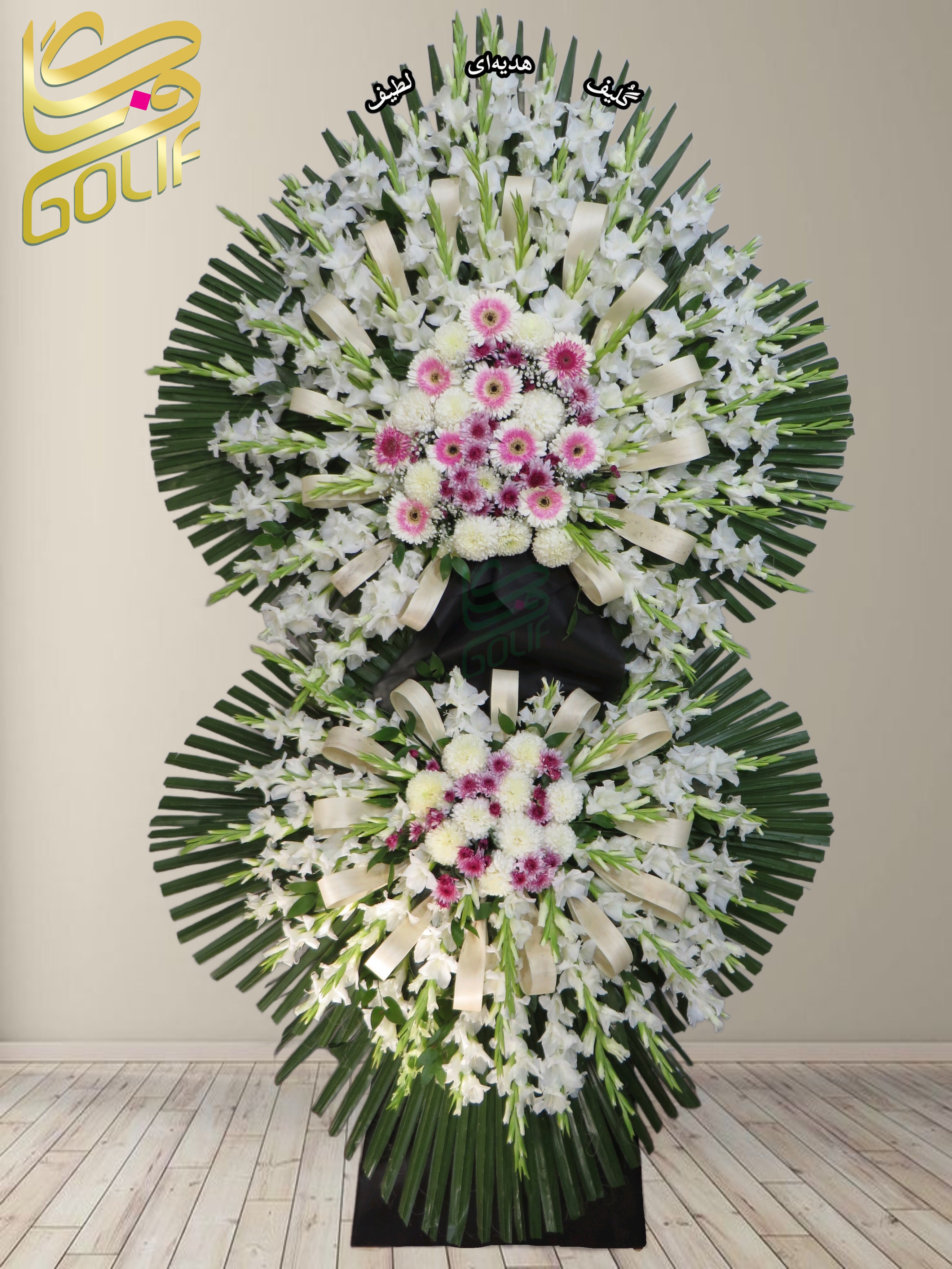 تاج گل تسلیت دو طبقه (گلایل، داوودی، رز هلندی) تاج گل ختم، تاج گل ترحیم، تاج گل مراسم ترحیم گل‌هایی که در طراحی این تاج گل دیده می‌شوند شامل موارد زیر هستند: 1. **گلایل سفید:** برجسته‌ترین گل که به نماد پاکی، همدردی و احترام شناخته می‌شود. 2. **گل داوودی سفید:** حضور قابل‌توجهی در طراحی دارد و نمایانگر صداقت و آرامش است. 3. **رز هلندی کرم با حاشیه صورتی:** نشانه‌ای از عشق و خاطرات ماندگار که جذابیت خاصی به ترکیب داده است. 4. **لیسیانتوس سفید:** با ظاهری لطیف که پیام همدلی و همراهی در غم و اندوه را منتقل می‌کند. 5. **برگ‌های تزئینی (مثل نخل):** طراحی را با رنگ سبز کامل کرده و باعث شکوه تاج گل شده است. 6. **روبان کرم یا سفید:** جلوه‌ای رسمی و زیبا به ترکیب اضافه کرده است. سفارش آنلاین تاج گل سفارش آنلاین گل عکاسی شده توسط گل‌فروشی آنلاین گلیف