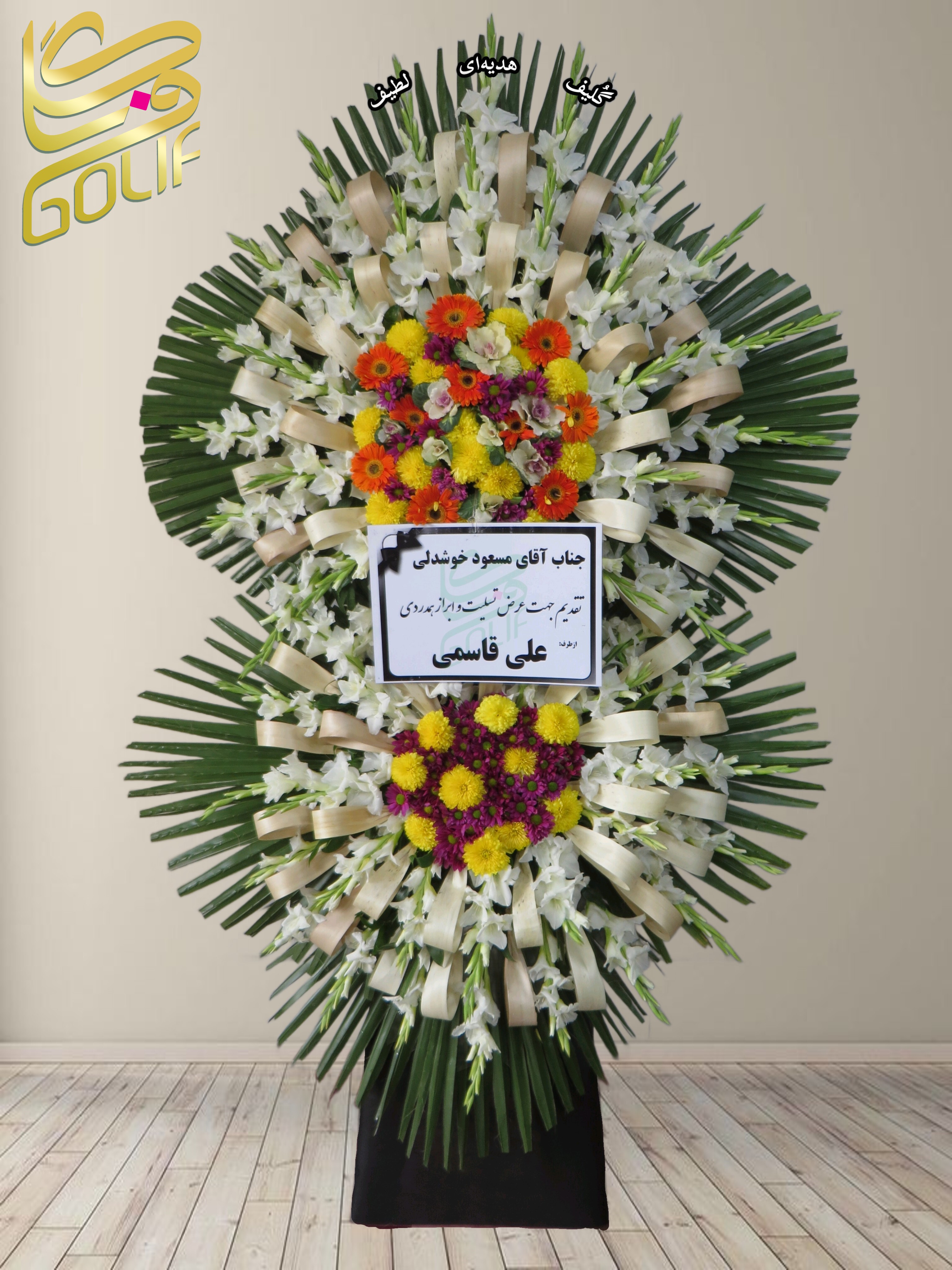 تاج گل تسلیت دو طبقه (گلایل، داوودی، رز هلندی) تاج گل ختم، تاج گل ترحیم، تاج گل مراسم ترحیم گل‌هایی که در طراحی این تاج گل دیده می‌شوند شامل موارد زیر هستند: گلایل سفید: که سمبل پاکی و احترام است و در لایه‌های مختلف طراحی حضور برجسته‌ای دارد. داوودی سفید: سمبل آرامش و صداقت، که در بخش‌های مختلف تاج گل دیده می‌شود. رز هلندی کرم با حاشیه صورتی: نمایانگر عشق و خاطرات ماندگار، که در طراحی رنگی جلوه‌ای خاص ایجاد کرده است. لیسیانتوس سفید: با ظاهری لطیف، حس همدلی و همراهی را تقویت می‌کند. برگ‌های تزئینی سبز: مانند نخل، که به طراحی حجم و جلوه خاصی بخشیده‌اند. روبان‌های کرم یا سفید: که تأکیدی بر رسمیت و زیبایی تاج گل است. سفارش آنلاین تاج گل سفارش آنلاین گل عکاسی شده توسط گل‌فروشی آنلاین گلیف