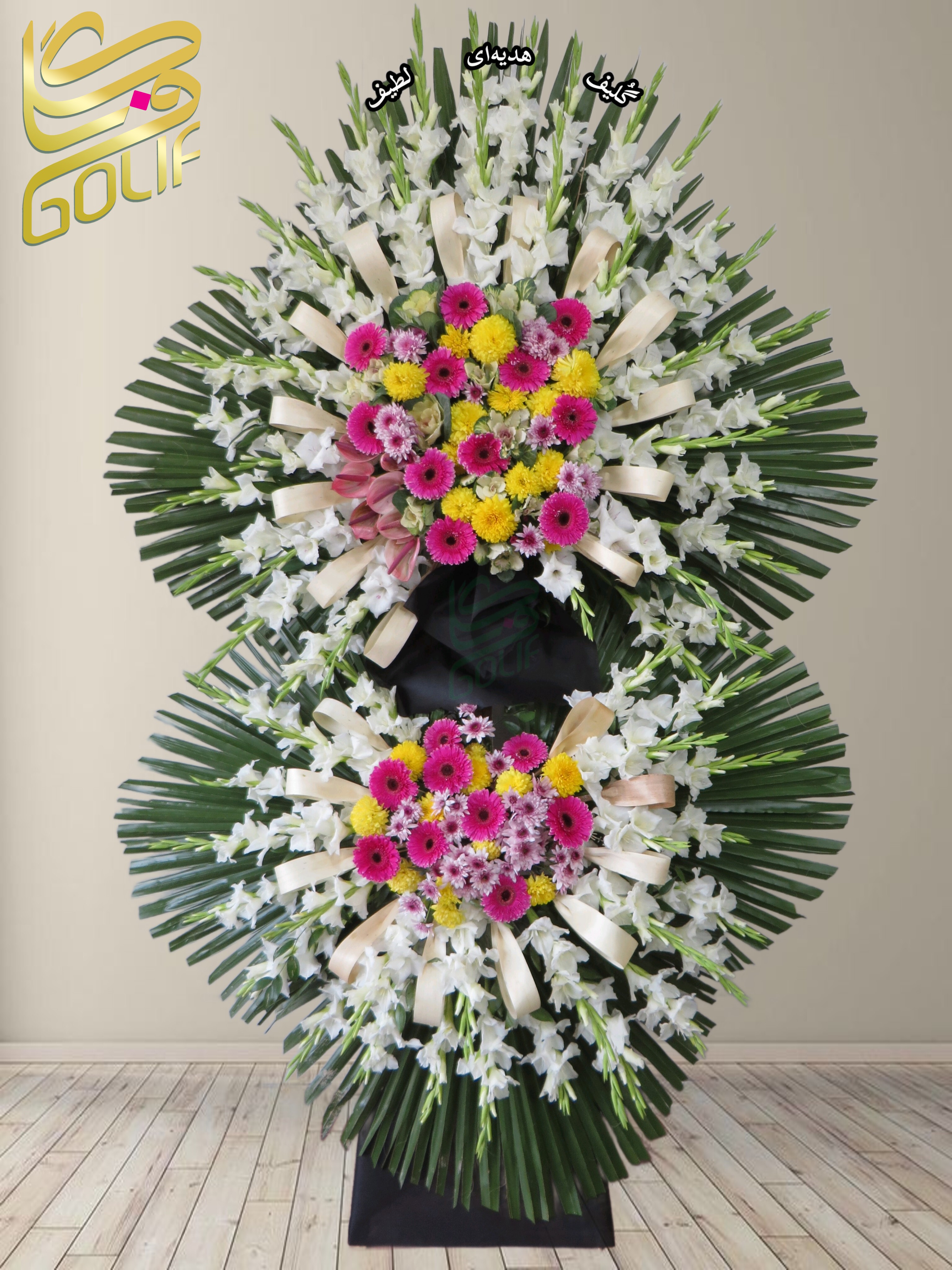 تاج گل تسلیت دو طبقه (گلایل، داوودی، رز هلندی صورتی) تاج گل ختم، تاج گل ترحیم، تاج گل مراسم ترحیم گل‌هایی که در طراحی این تاج گل دیده می‌شوند شامل موارد زیر هستند: ✅ گلایل سفید: که نماد پاکی و احترام است و در طراحی این تاج گل برجسته است. ✅ داوودی سفید: حضور چشمگیری دارد و حس صداقت و آرامش را به ترکیب القا می‌کند. ✅ رز هلندی کرم با حاشیه صورتی: که یادآور عشق و خاطرات ماندگار است و به طراحی تاج گل جلوه‌ای ملایم و متعادل می‌دهد. ✅ لیسیانتوس سفید: با بافت لطیف خود، حالتی از همدلی و همراهی را منتقل می‌کند. ✅ برگ‌های تزئینی سبز مانند نخل: که علاوه بر افزایش شکوه ترکیب، رنگ سبز طبیعی را به طراحی افزوده‌اند. ✅ روبان‌های سفید یا کرم: که نمایی رسمی و زیبا را به ترکیب این تاج گل بخشیده‌اند. ترکیب این عناصر و گل‌ها نه‌تنها زیبایی، بلکه پیام تسلیت و احترام خاصی را به‌خوبی منتقل می‌کند. سفارش آنلاین تاج گل سفارش آنلاین گل عکاسی شده توسط گل‌فروشی آنلاین گلیف