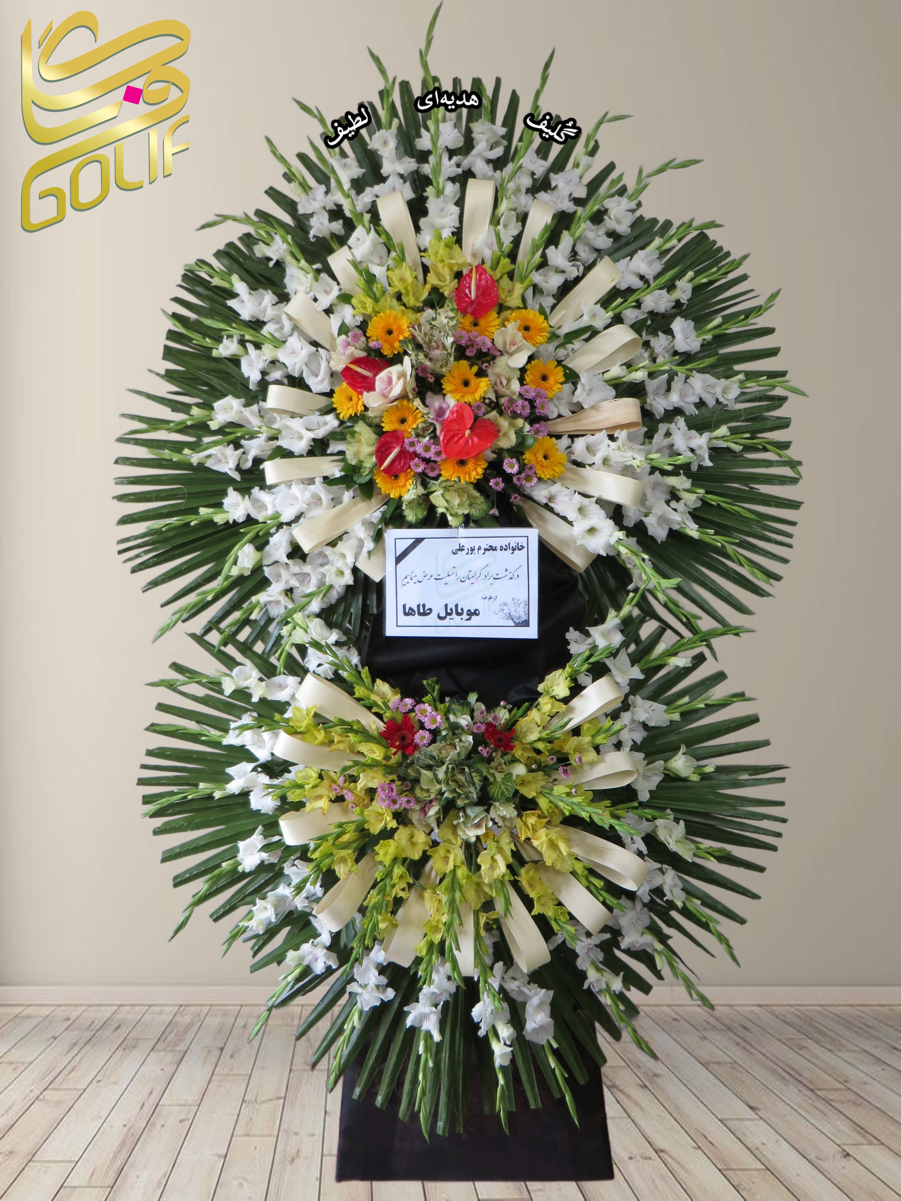 تاج گل تسلیت دو طبقه (رز، داوودی، لیلیوم، میخک) تاج گل ختم، تاج گل ترحیم، تاج گل مراسم ترحیم گل‌هایی که در طراحی این تاج گل دیده می‌شوند شامل موارد زیر هستند: تصویر شامل یک دسته گل بزرگ و زیبا است که به شکل دو دایره روی هم چیده شده است. هر بخش از گل‌ها به صورت متقارن طراحی شده و ترکیبی از گل‌های سفید، زرد، قرمز، صورتی و بنفش دارد. میان این گل‌ها برگ‌ها و روبان‌ها به کار رفته‌اند که به زیبایی دسته گل افزوده‌اند. گل‌های رز: رنگ‌های مختلفی مثل سفید، صورتی و قرمز. گل‌های داوودی: معمولاً برای مراسم تسلیت استفاده می‌شوند. لیلیوم (سوسن): گل‌های بزرگ و خوشبو که ممکن است سفید یا صورتی باشند. میخک: گل‌هایی با رنگ‌های متنوع که اغلب در ترکیب با گل‌های دیگر دیده می‌شوند. این گل‌ها برای یک مراسم تسلیت ارسال شده باشند، چون بر روی کارت وسط دسته گل، پیامی به زبان فارسی برای خانواده پور علی نوشته شده است که ابراز همدردی و تسلیت می‌کند. سفارش آنلاین تاج گل سفارش آنلاین گل عکاسی شده توسط گل‌فروشی آنلاین گلیف