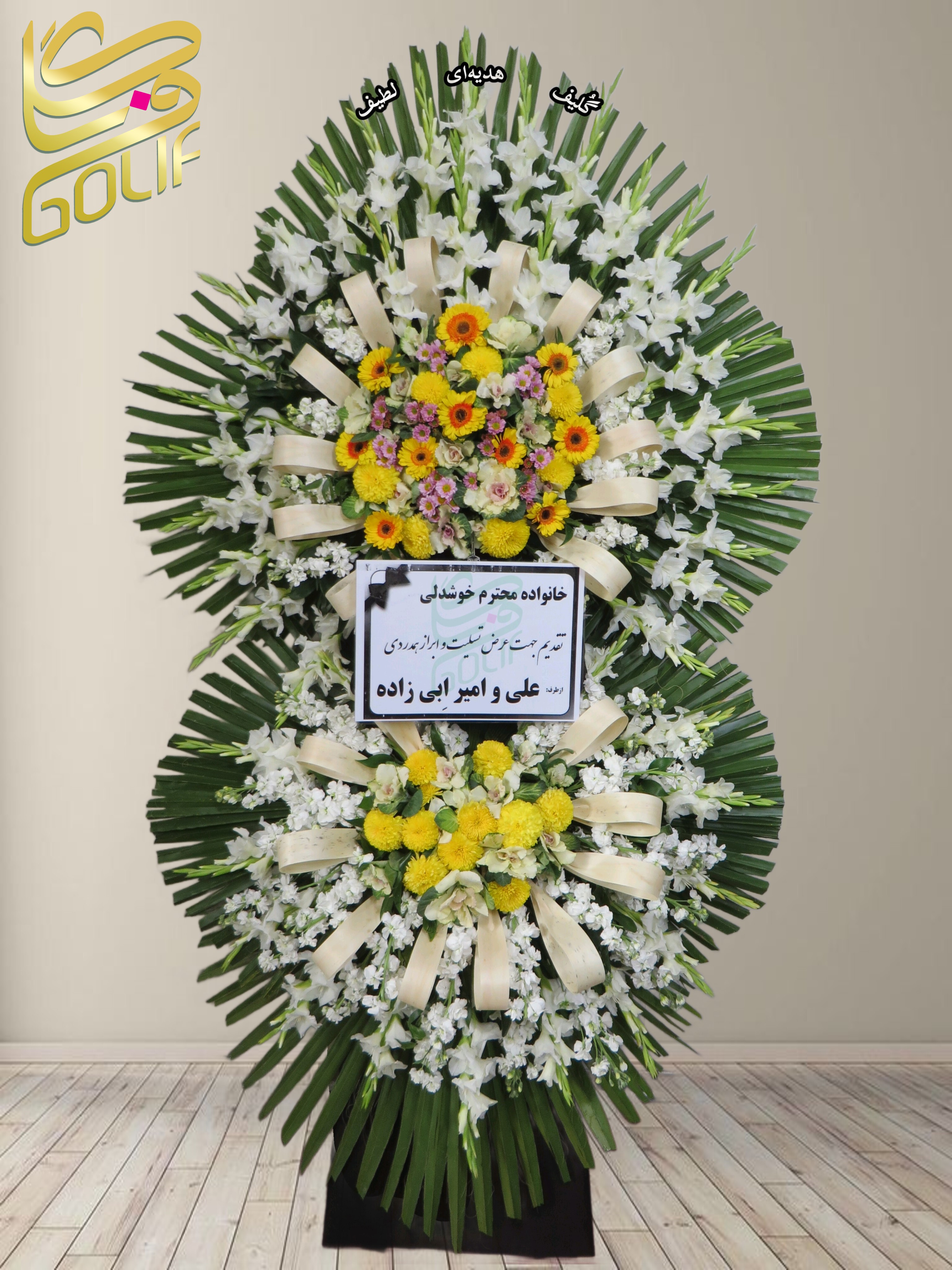 تاج گل تسلیت دو طبقه (لیلیوم، داوودی، میخک) تاج گل ختم، تاج گل ترحیم، تاج گل مراسم ترحیم گل‌هایی که در این تاج گل دیده می‌شوند شامل موارد زیر هستند: سوسن (لیلیوم سفید): گل‌هایی بزرگ و زیبا با عطر خوش، که به وضوح در این تصویر مشخص هستند. داوودی (کرزنتیم): گل‌های زرد رنگ که برای مراسم تسلیت بسیار معمول هستند. میخک‌های رنگارنگ: به نظر می‌رسد تعدادی گل‌ میخک در این دسته گل به کار رفته باشد. این ترکیب گل‌ها با برگ‌های سبز و روبان‌های کرم رنگ بسیار زیبا طراحی شده است. سفارش آنلاین تاج گل سفارش آنلاین گل عکاسی شده توسط گل‌فروشی آنلاین گلیف